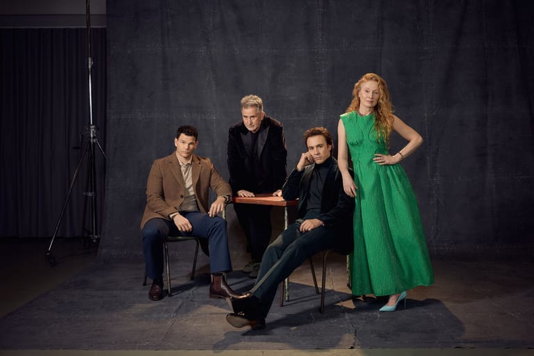 Four people pose together against a dark backdrop, three seated and one standing in a green dress.