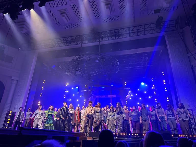 Large ensemble cast stands on stage bathed in blue lighting and dramatic spotlights during a theatre production.