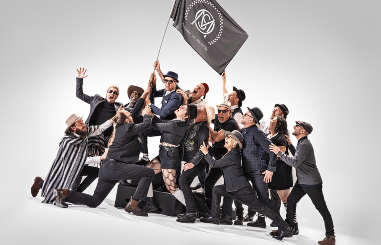 Large group of people in dark clothing clustered together, one holding aloft a dark flag with a circular emblem.