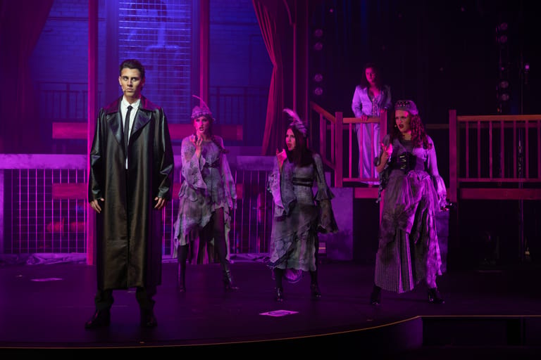 Young performers in tattered costumes stand on a stage bathed in purple lighting during Hadestown: Teen Edition.
