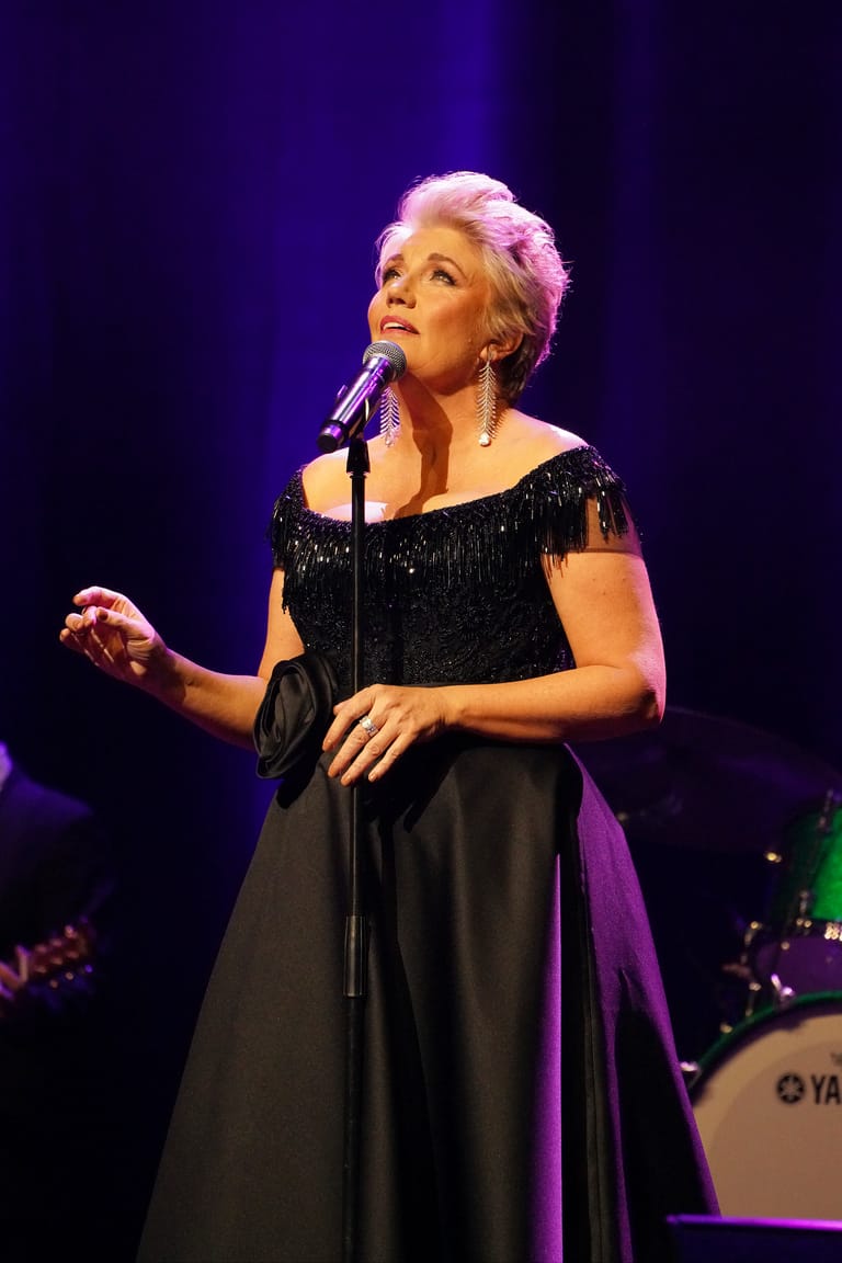 Performer with short blonde hair singing at a microphone in a black sequined top under purple stage lighting.