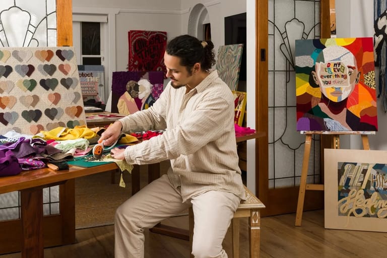 Artist in cream-coloured studio clothing arranging colourful fabric and materials at a wooden work table.