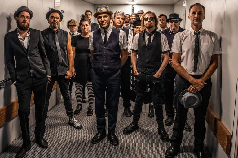 Large ensemble of performers in formal attire standing together in a backstage corridor.