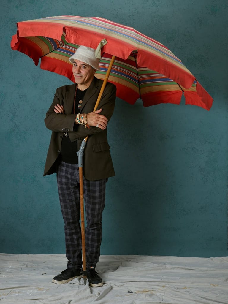 Person in dark jacket and patterned trousers holding a large red and striped umbrella against a teal background.