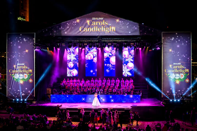 Large stage production with performer in white gown centre stage, surrounded by ensemble cast and dramatic purple and blue lighting, ornate banner overhead.