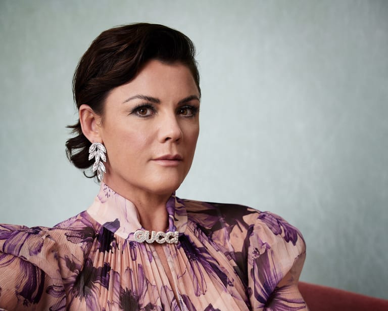 Woman with dark hair in purple patterned top and silver earrings against neutral grey background.