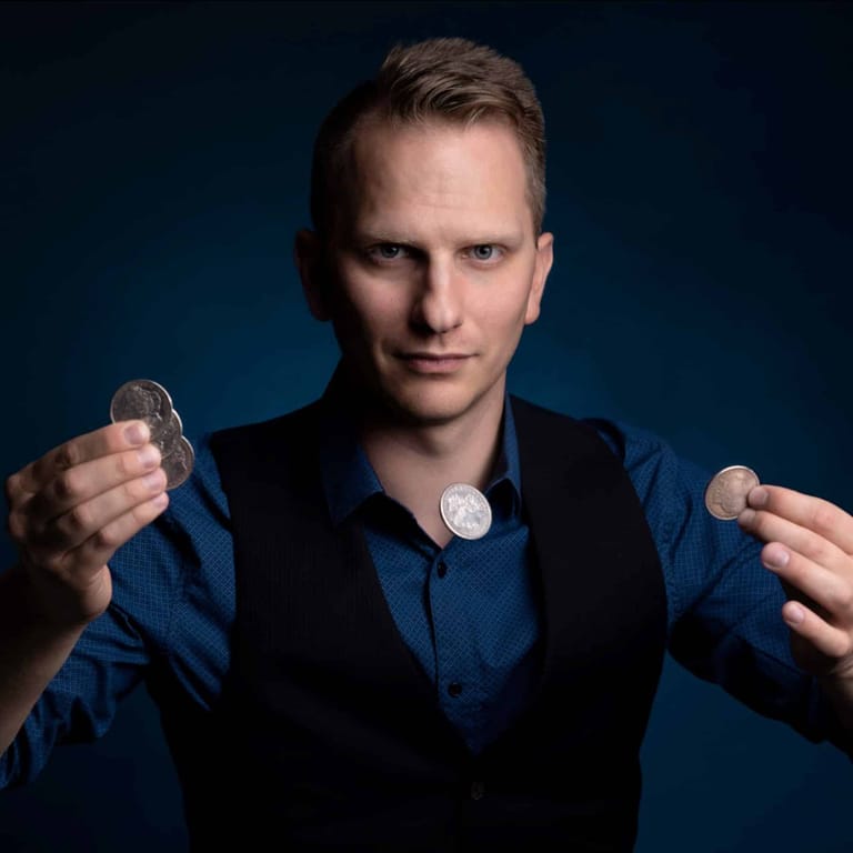 Magician in dark blue shirt holding coins in both hands against a blue backdrop.