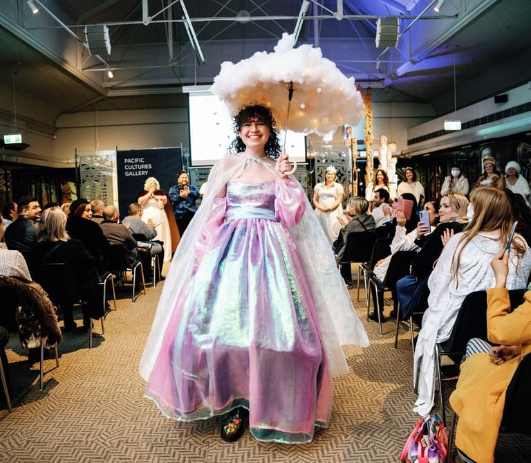 Model wearing an elaborate pink and pastel gown with voluminous white cloud-like headpiece walks a runway before seated audience members.