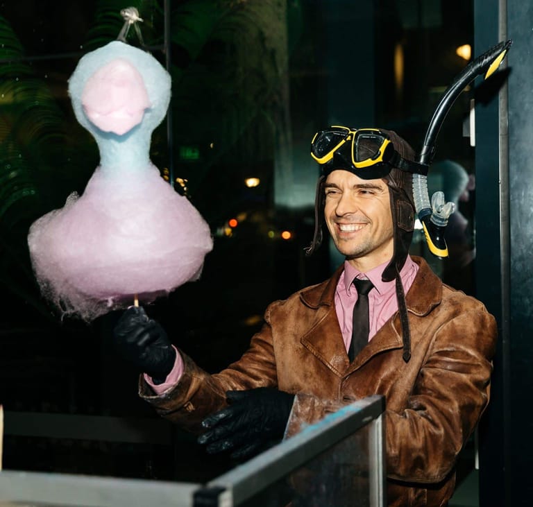 Man in brown jacket and tricorn hat holds oversized fluffy white puppet head at night outdoors.