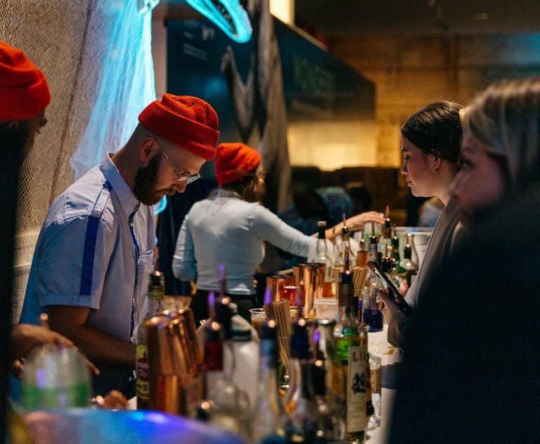 Bartender in red cap pouring drinks at a dimly lit bar counter for patrons.