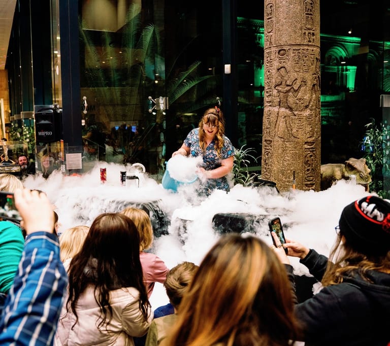 Performer surrounded by theatrical fog sits on stage amid spectators holding phones at a night event.