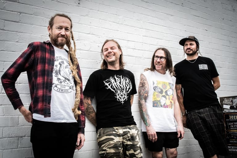 Four band members stand smiling against a white brick wall, wearing casual graphic t-shirts and patterned bottoms.