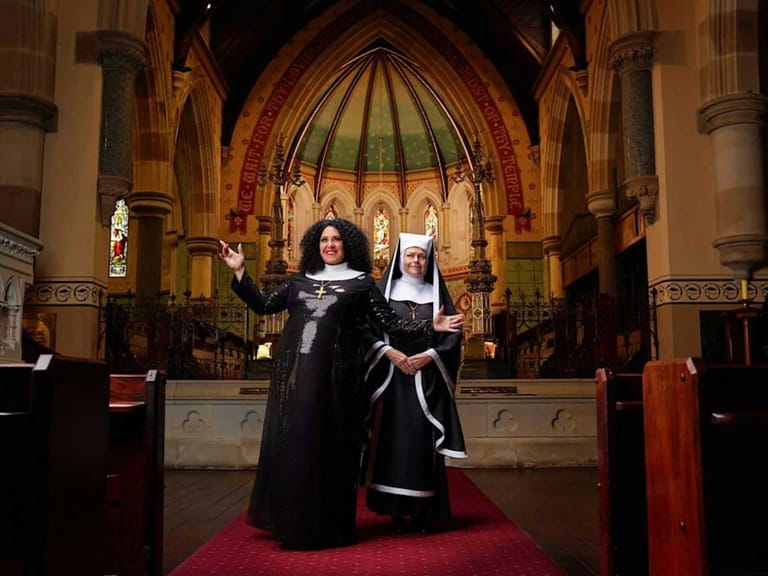 Two performers in nun habits stand on the altar of a grand gothic church interior lit by stained glass windows.