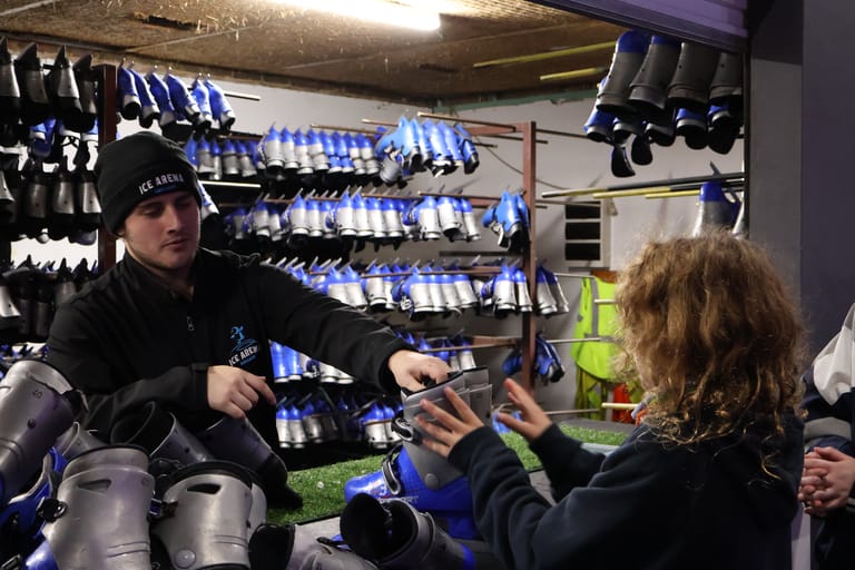 Technician in black beanie fits equipment to performer's arm backstage near shelves of blue lighting gear.