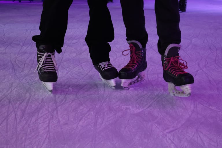 Four people wearing ice skates stand on a purple-lit ice rink.