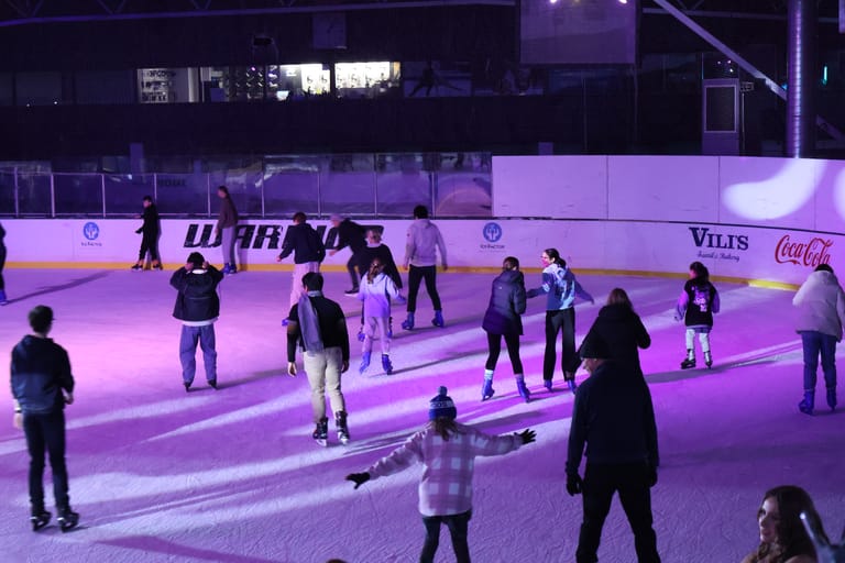 Skaters in purple-lit ice rink with spectators and advertising boards visible in background.