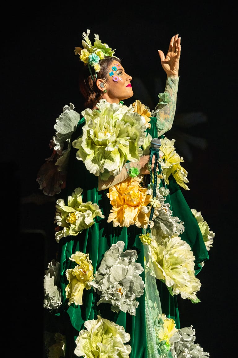 Performer in elaborate floral costume of oversized yellow and white flowers with green dress, arm raised against dark stage backdrop.