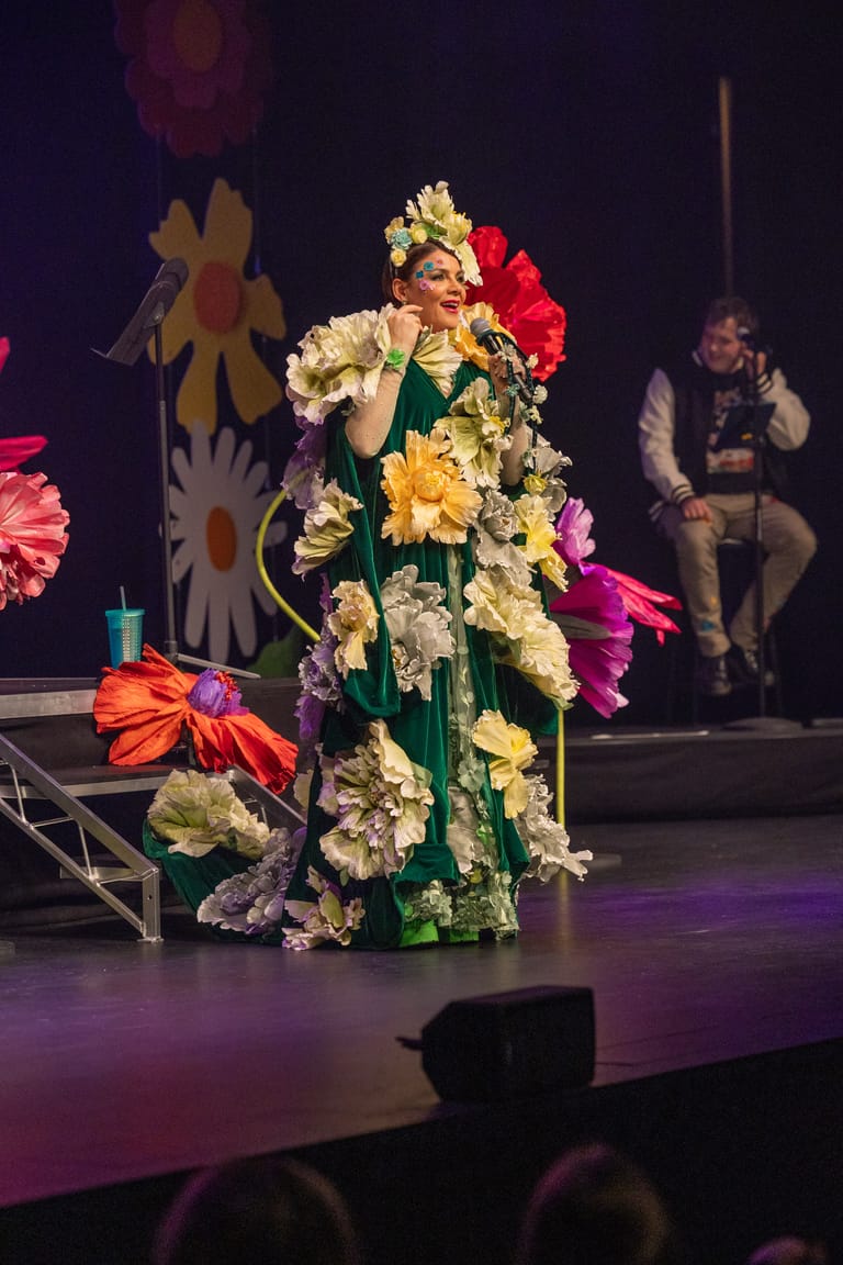 Performer in a green dress covered with oversized colourful flowers stands centre stage under purple lighting.