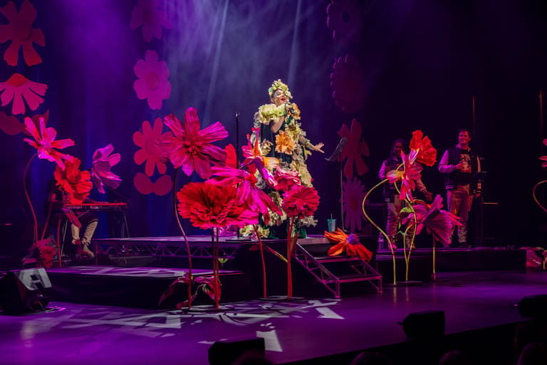 Performer in gold outfit centre stage surrounded by large illuminated red flowers under purple stage lighting.