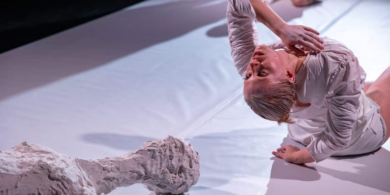 Performer in grey clothing lies on a white stage floor beside sculptural props during a contemporary theatre production.