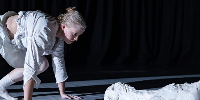 Performer in white shirt crouched on stage beside flowing white fabric against dark backdrop.