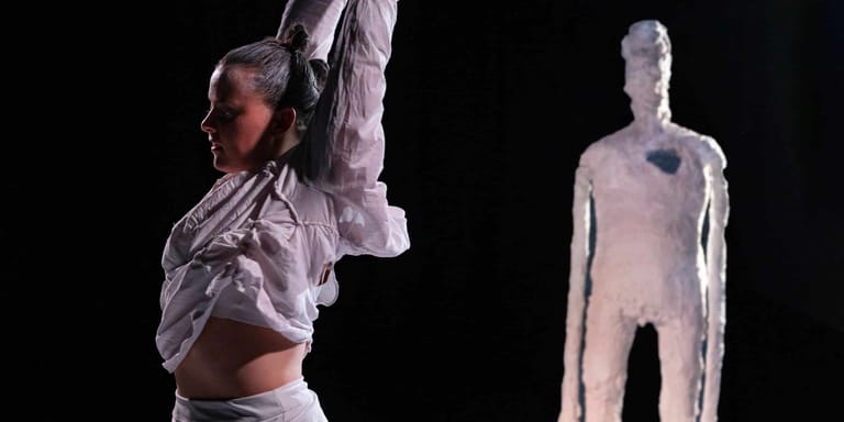 Performer in white clothing executing an overhead stretch pose whilst another figure stands in the background under stage lighting.
