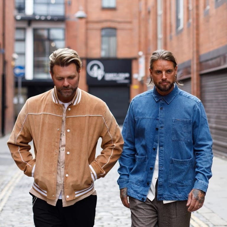 Two men in casual denim and suede jackets walk side-by-side down a brick-paved urban lane lined with heritage buildings.