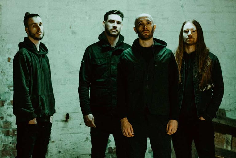 Four band members stand against a weathered mint-green wall in dark clothing.