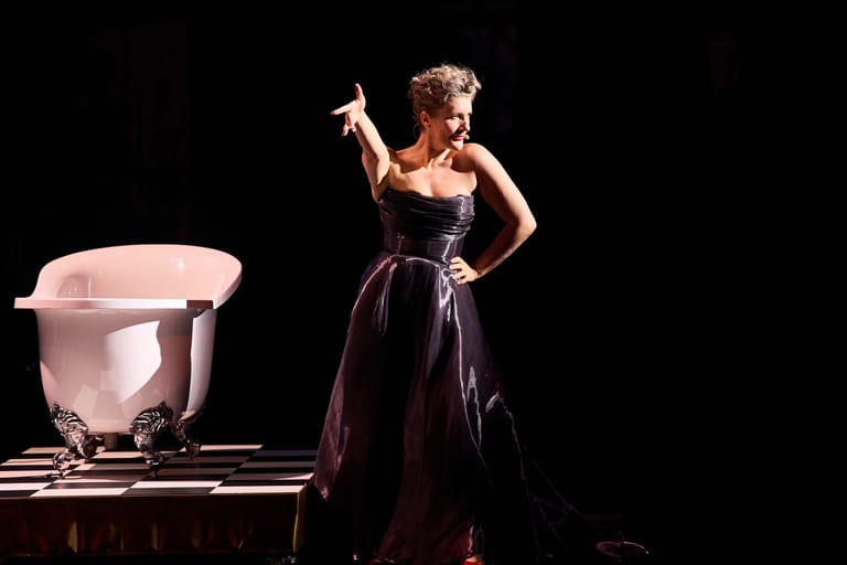 Performer in a dark evening gown gestures expressively beside a large pink sculptural object on a theatrical stage.