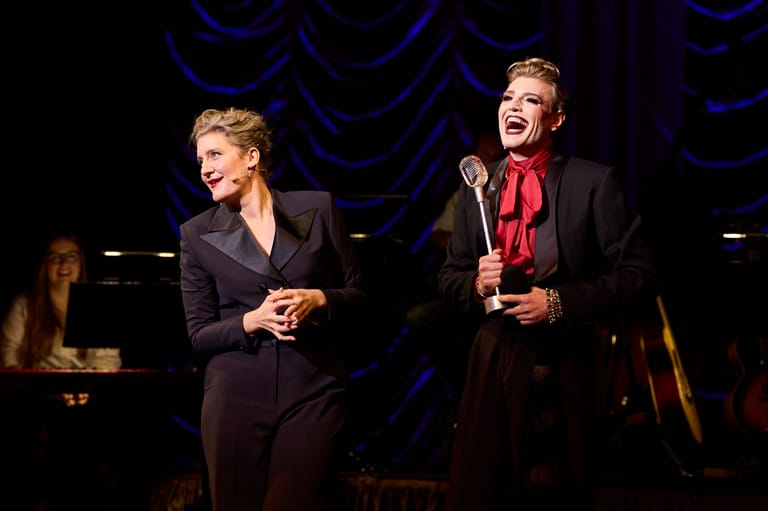 Two performers in dark suits with red accents stand centre stage under blue theatrical lighting, holding microphones during a live performance.