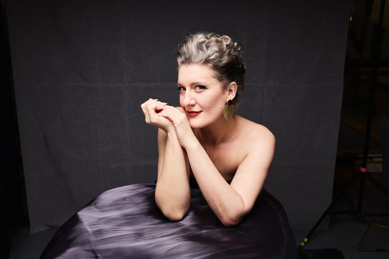 Woman with styled blonde hair and red lipstick posed against dark background, resting chin on clasped hands.
