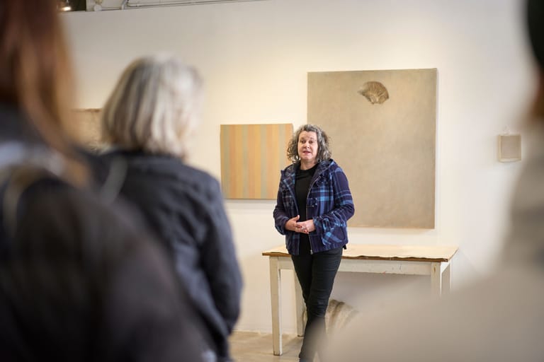 Person in blue jacket speaking to audience members in gallery space with artworks on beige walls.