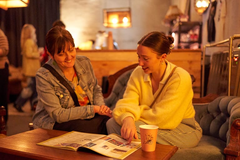 Two women in casual clothing examine a magazine together at a wooden table in a warm, lamp-lit interior space.