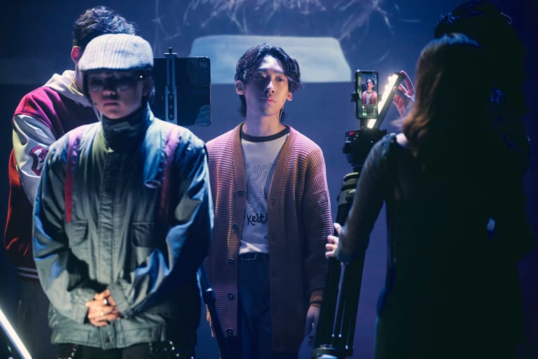 Young musicians on stage conversing beneath blue lighting, with audience members visible in the background.
