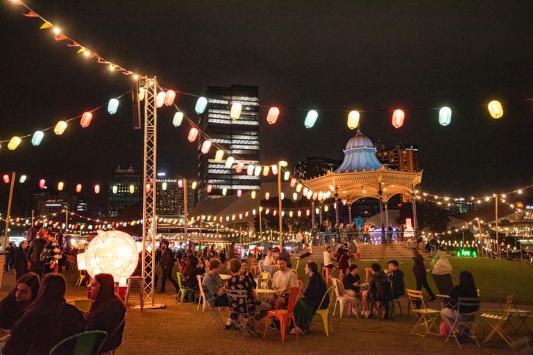 Festive outdoor night gathering with crowds under string lights and lanterns, illuminated marquee structures visible across the dark field.