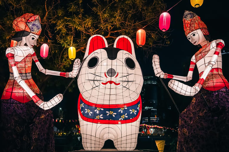 Large illuminated Maneki-neko (lucky cat) lantern flanked by two costumed figures holding coloured lanterns at night.