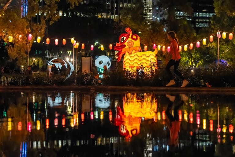 Illuminated lantern installations and projections reflect in still water at night, with city buildings glowing in the background.