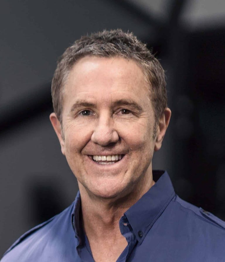 Man in navy blue shirt smiling at camera against blurred grey background.