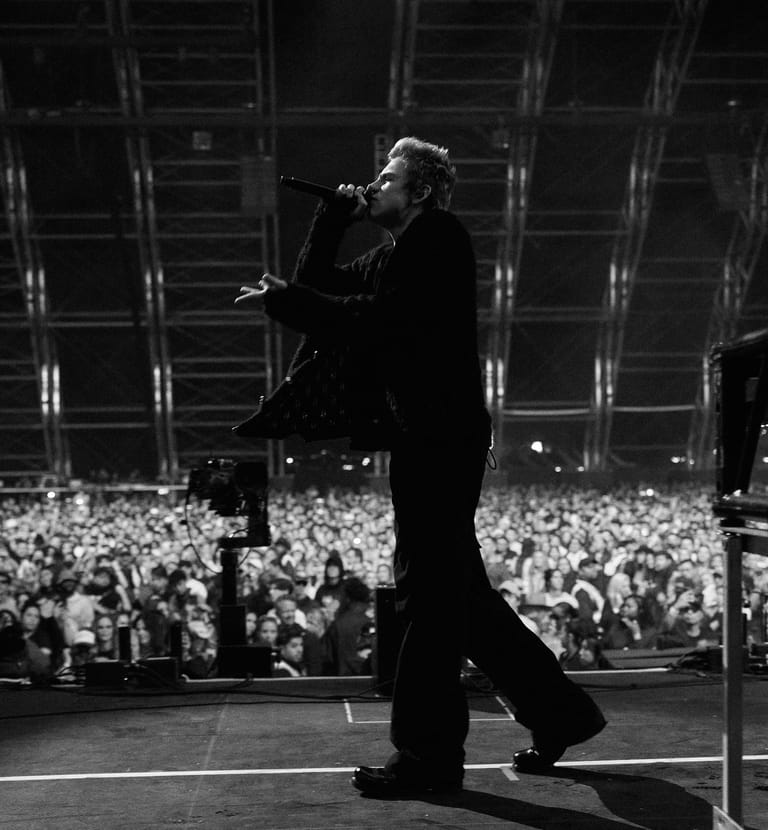 Singer in dark suit performing at a microphone before a large crowd in a stadium venue.