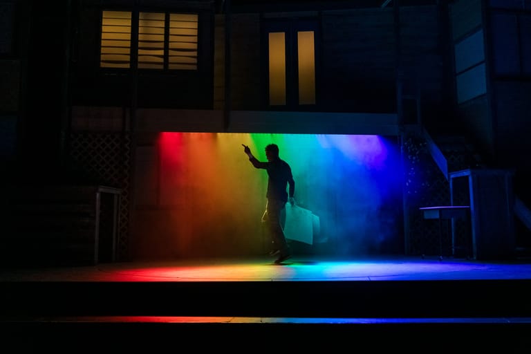 Silhouetted performer stands on stage bathed in rainbow-coloured lighting against a dark backdrop.