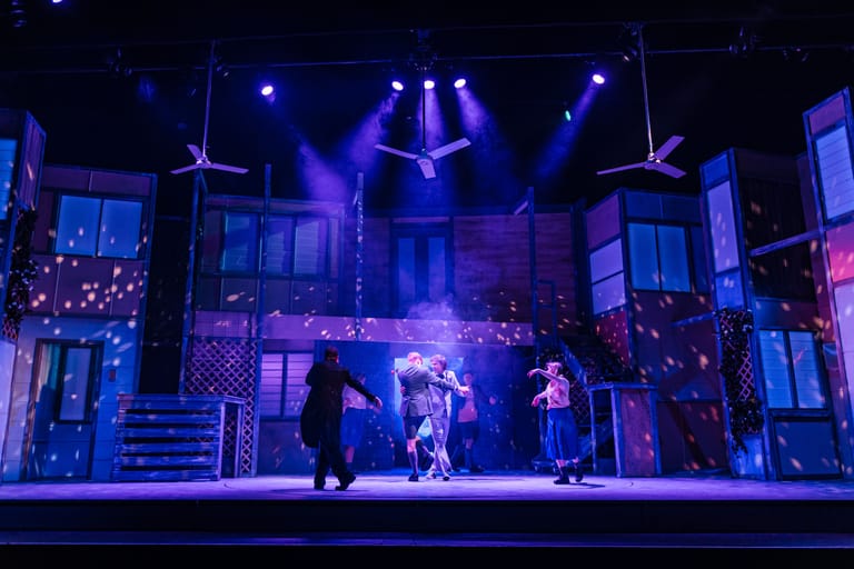 Performers on a blue and purple lit stage set with large window props and overhead lighting rigs.