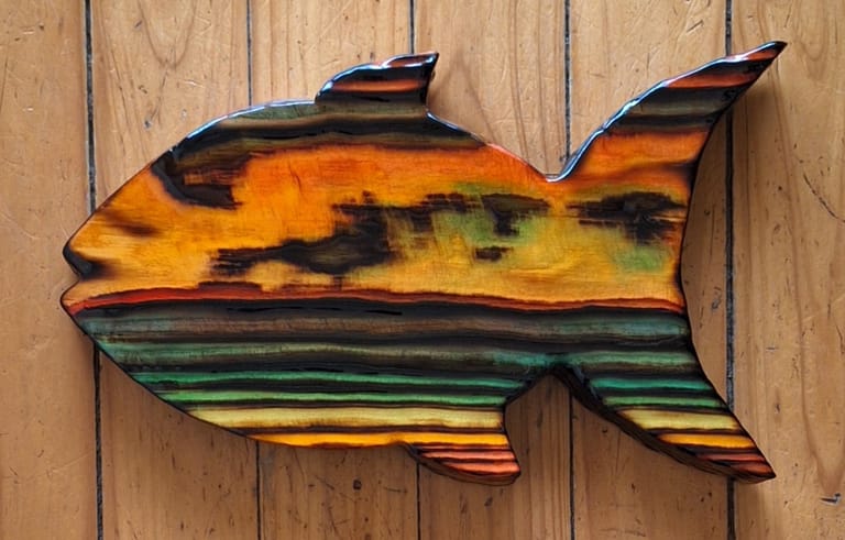 Colourful wooden fish sculpture with orange, yellow, green and black stripes mounted on timber wall.