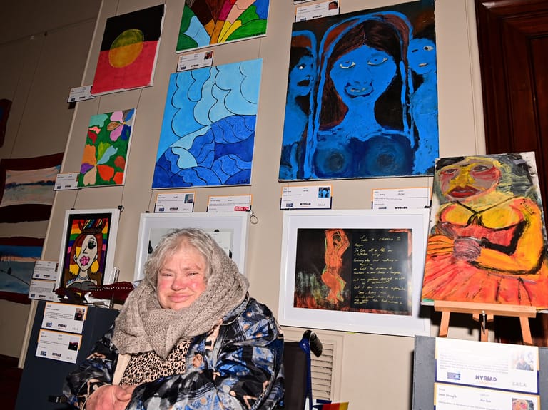 Artist stands before a gallery wall displaying colourful portrait paintings and mixed-media artworks in blues, oranges and vibrant hues.