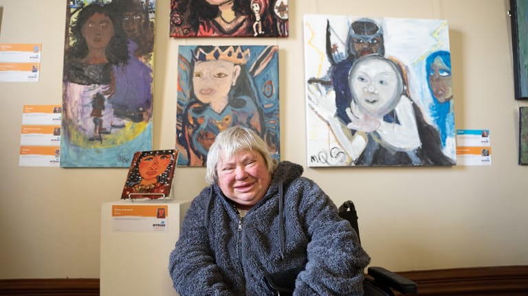 Woman with blonde hair seated in front of displayed artworks featuring painted portraits on a gallery wall.