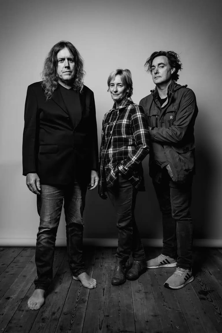Three musicians in dark casual attire stand barefoot against a grey wall in black and white portrait.