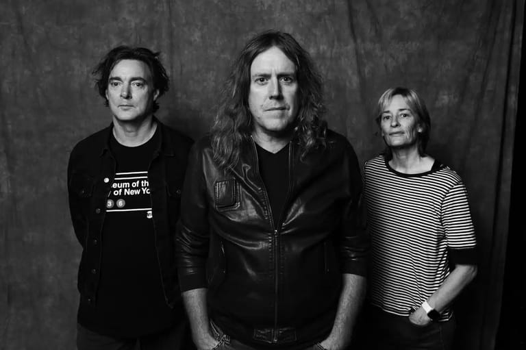 Three musicians pose against a grey backdrop in black and white portrait photography.