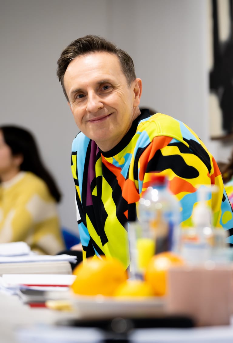 Man in a vibrant multicoloured geometric jacket smiles at a table during an event or discussion.