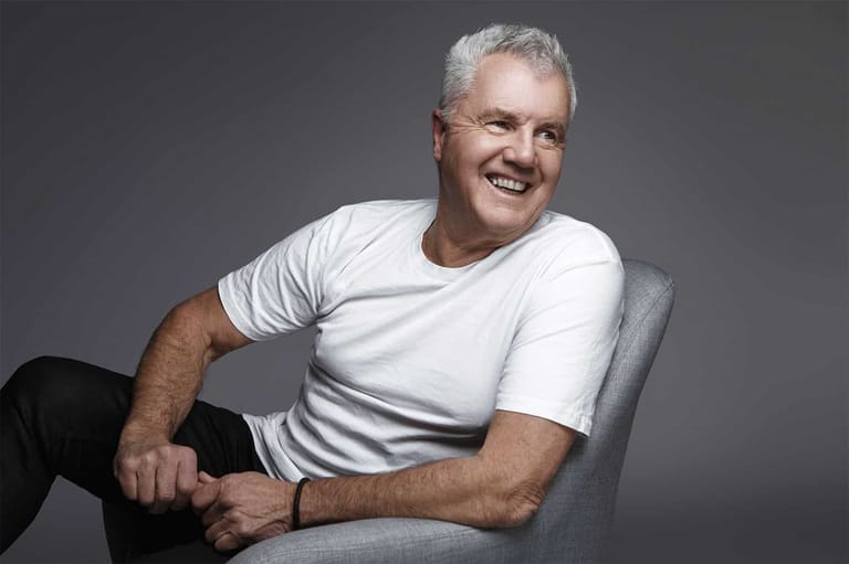 Middle-aged man with grey hair smiling at camera, wearing white t-shirt seated against grey background.