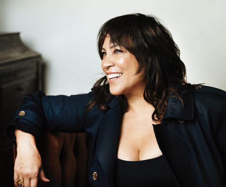 Woman with dark wavy hair smiling, wearing a navy blazer over a black top, seated indoors.