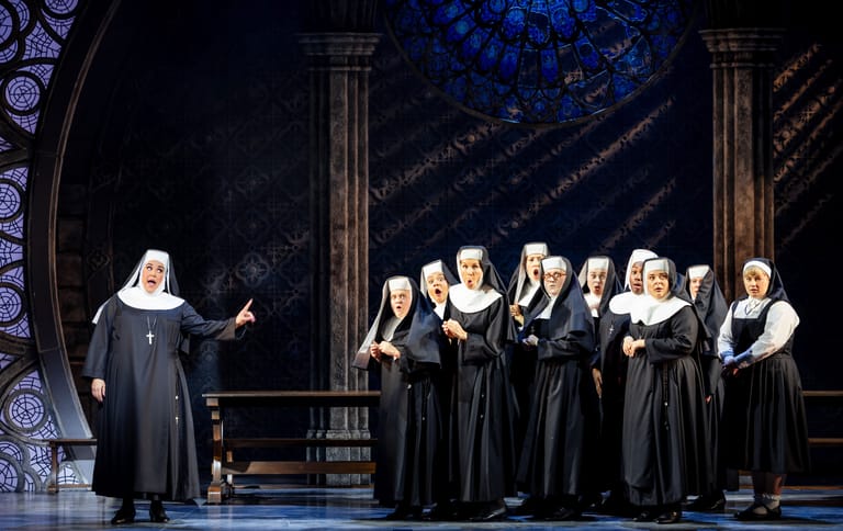 Choir of nuns in black habits singing together on stage beneath ornate architectural columns.